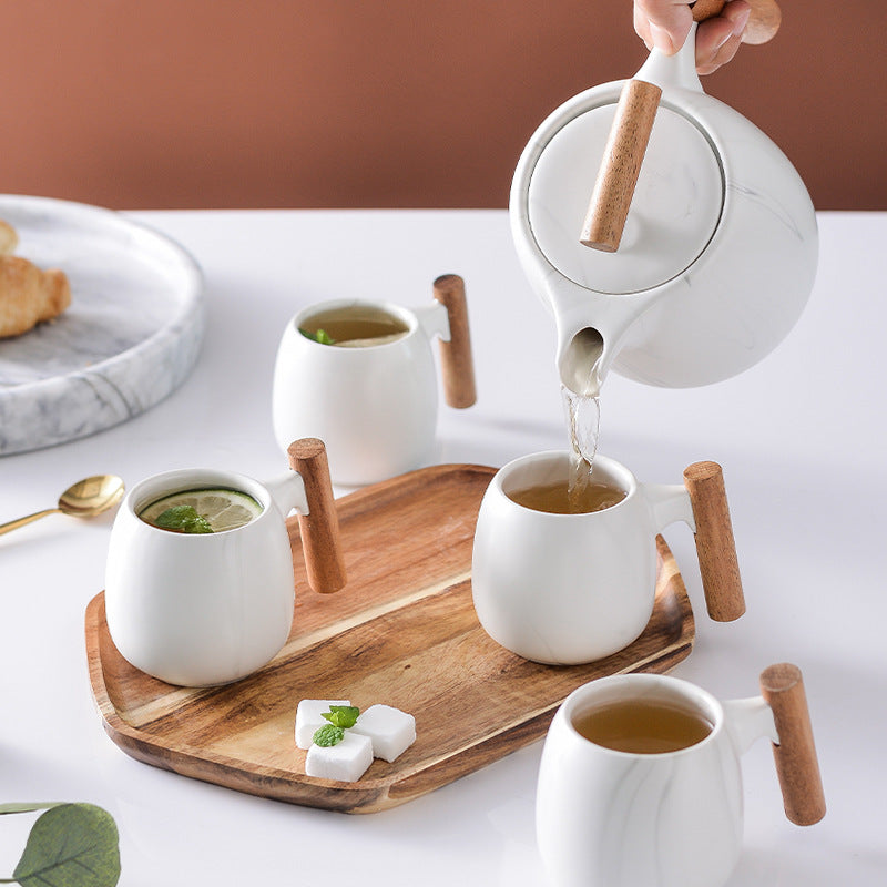 A white ceramic tea set with light wood handles and spout covers is arranged on a wooden tray, with one teapot being poured into two cups. A hand is visible holding the teapot, and the scene includes a white table, a brown wall, a marble plate with a pastry, and small garnish details.