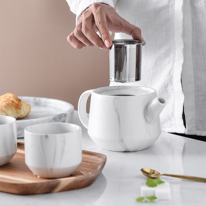 A person in a white shirt holds a stainless steel infuser above a white marble teapot on a white table. Two matching white marble cups sit on a wooden tray, with a plate holding a croissant in the background.