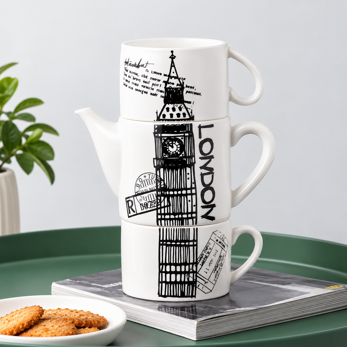Three stacked white ceramic mugs with black London-themed illustrations, including Big Ben, the word 'LONDON,' stamps, and handwritten text, displayed on a green tray with a magazine and cookies nearby.