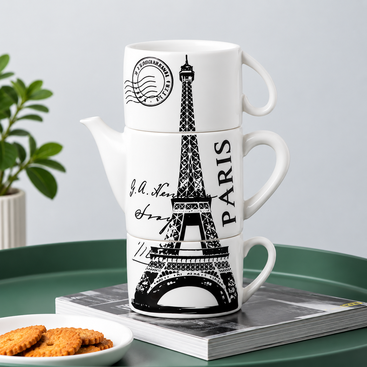 Three stacked white ceramic mugs with black Eiffel Tower and Paris-themed printed designs, styled on a green tray with a book and cookies in the background.