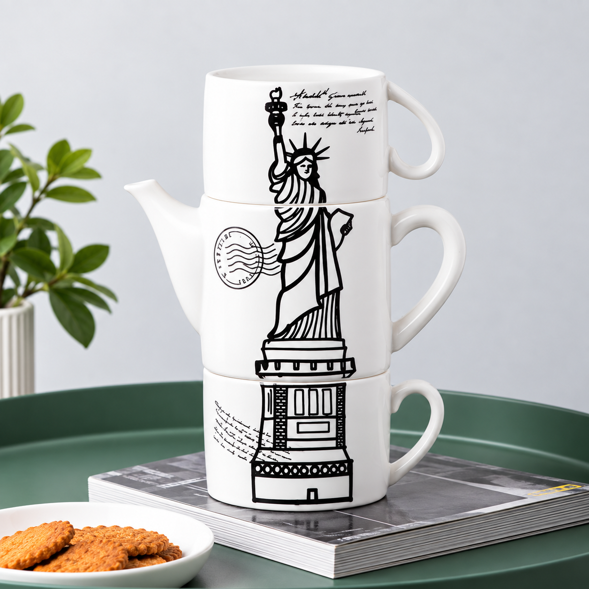 A stack of three white ceramic mugs with a black line-art illustration of the Statue of Liberty and surrounding architectural details, styled on a green tray with a book and cookies.
