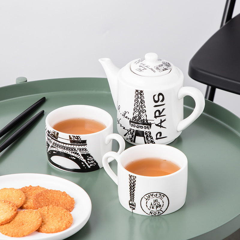 A white ceramic tea set with black Eiffel Tower illustrations and 'PARIS' text, arranged on a green tray with two cups of tea, chopsticks, cookies, and a chair edge visible.
