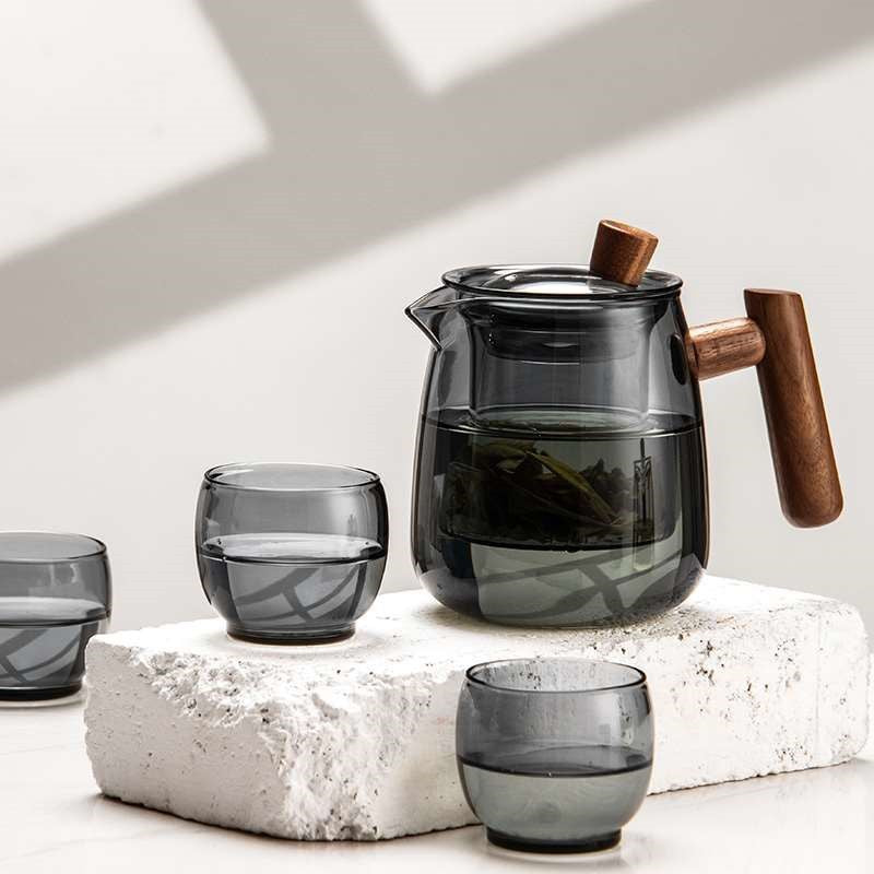 A dark smoky gray glass teapot with wooden handles and matching small glass cups, arranged on a white textured stone block against a softly lit neutral background.