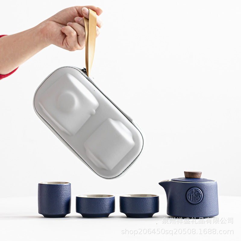 A hand holds a light gray rectangular organizer pouch with a tan handle above a matching dark blue ceramic tea set on a clean white background.