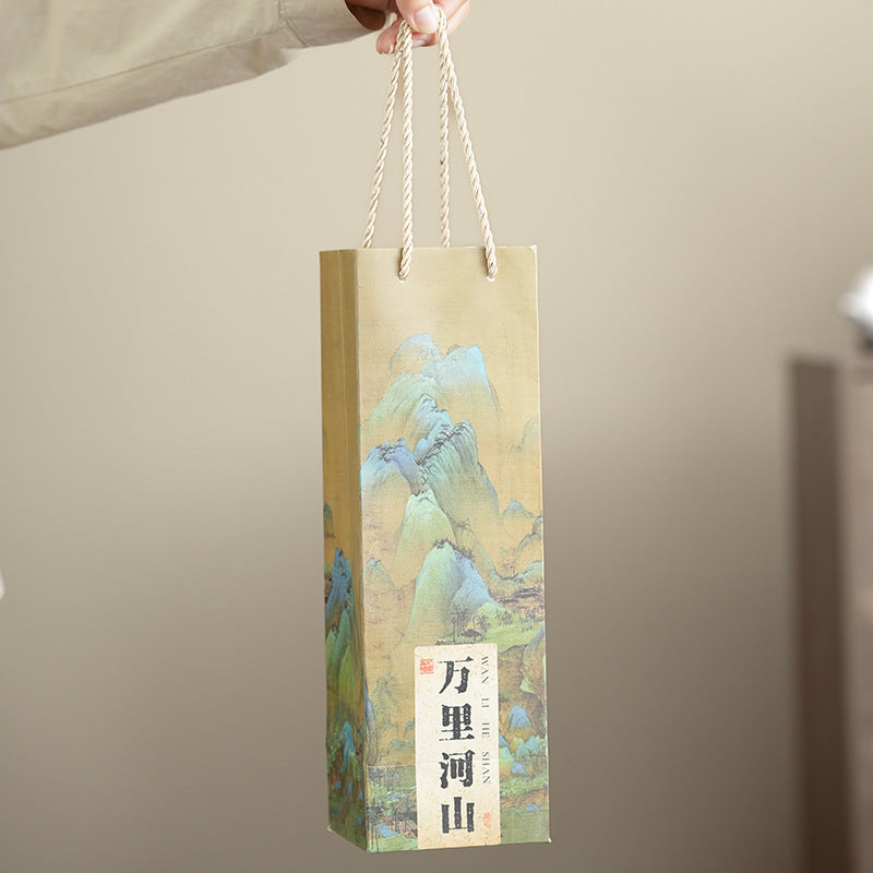 A hand holds a tall decorative paper gift bag with rope handles. The bag has a beige background with a printed landscape illustration in soft blue, green, and yellow tones, plus a vertical label with Chinese characters near the bottom.