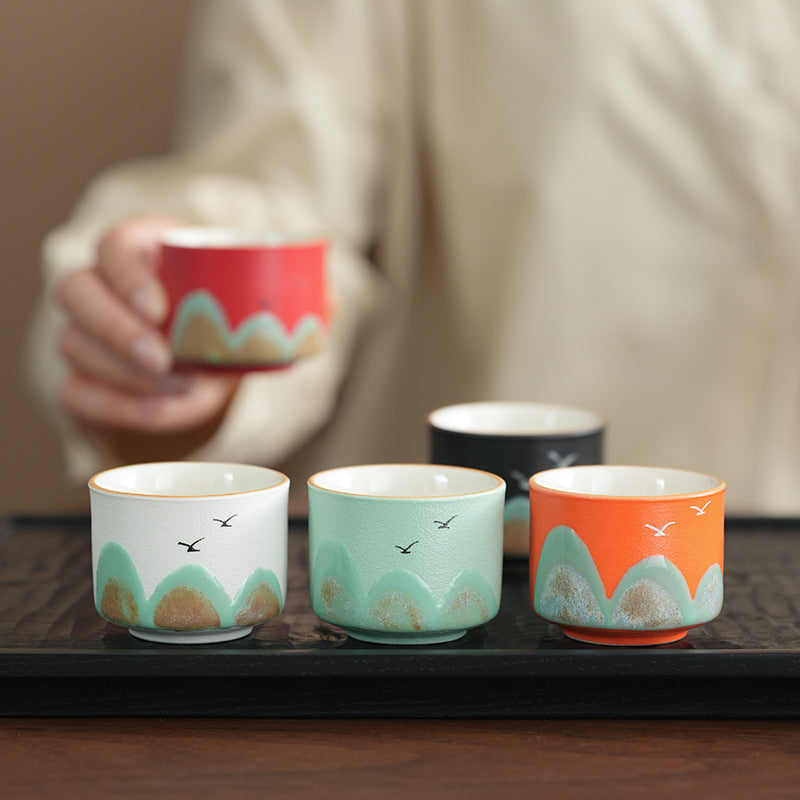 Four ceramic tea cups with hand-painted mountain and bird designs are arranged on a dark tray, with a person's hand holding a red cup in the blurred background.