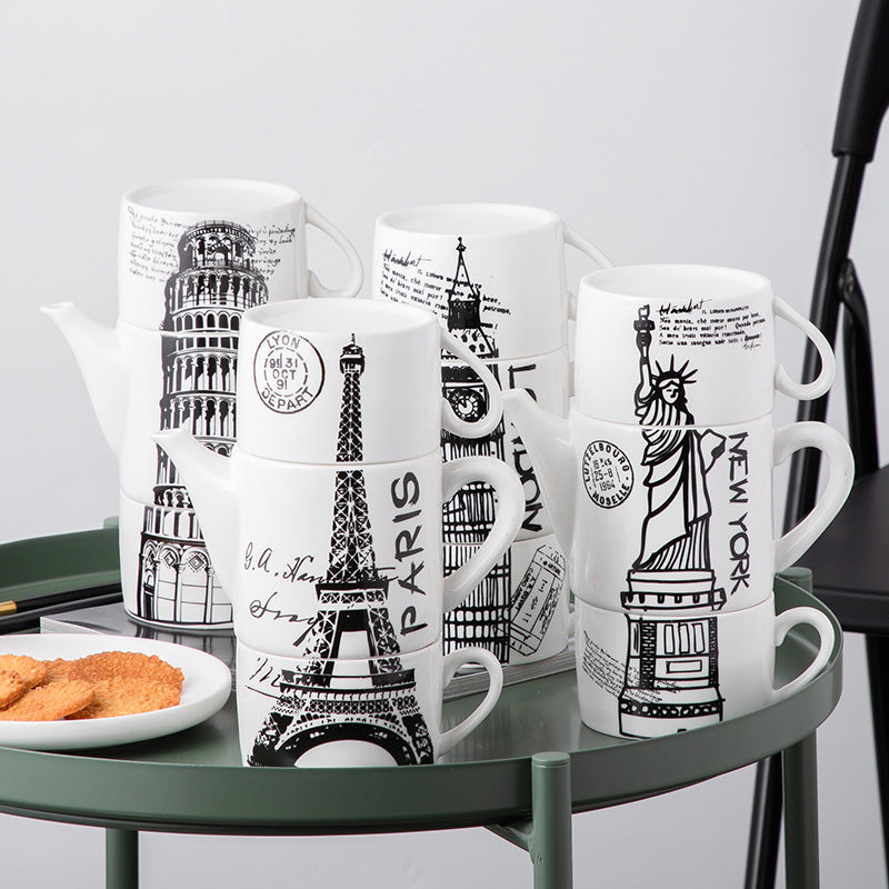 A set of white ceramic mugs and a matching teapot printed with black illustrations of famous landmarks, including the Leaning Tower of Pisa, Eiffel Tower, Big Ben, Statue of Liberty, and Taj Mahal, arranged on a green metal tray.