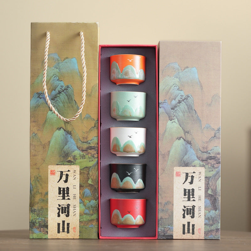 A ceramic tea set gift set displayed in its packaging, featuring four small cups with a multicolor landscape print, flanked by two tall decorative boxes with matching artwork and Chinese text.