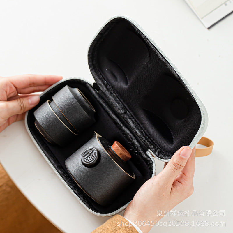 A black hard-shell carrying case with a black padded interior, shown open and held by two hands. Inside the case are two dark ceramic cups with light brown handles and a round decorative emblem on one cup.