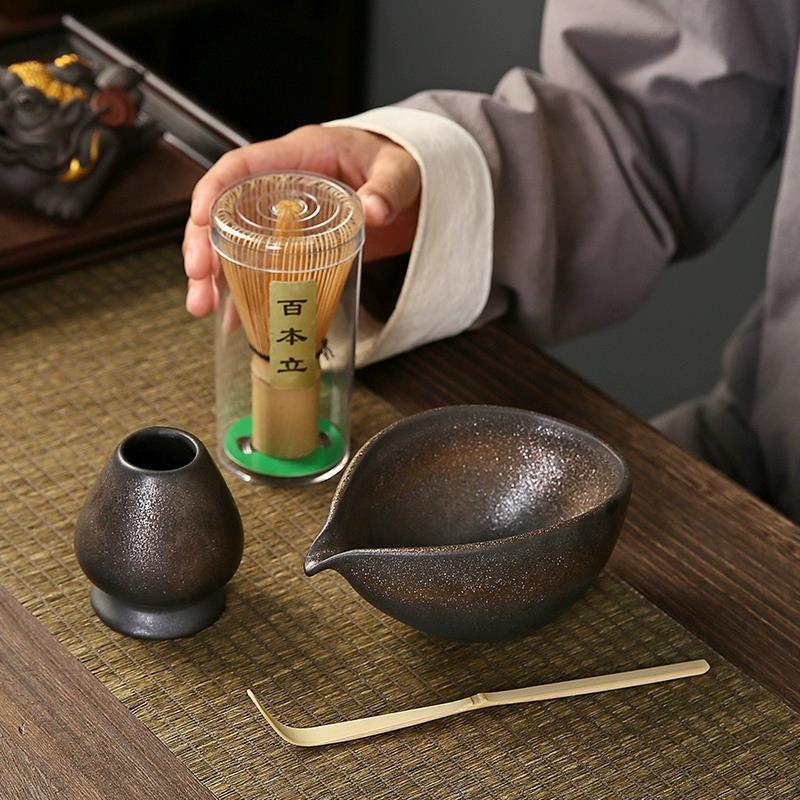 Person holding a glass container with tea leaves next to a black ceramic teapot and cup on a wooden table.