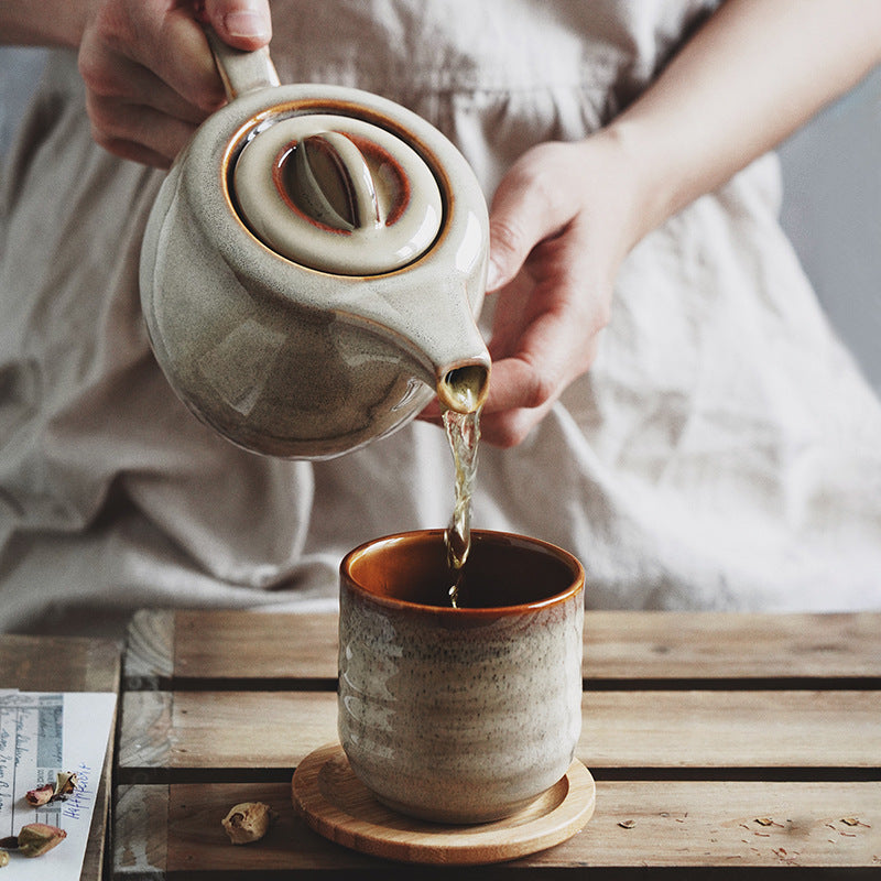A person in a beige dress pours tea from a speckled beige ceramic teapot into a matching speckled cup with a brown interior, set on a wooden table.