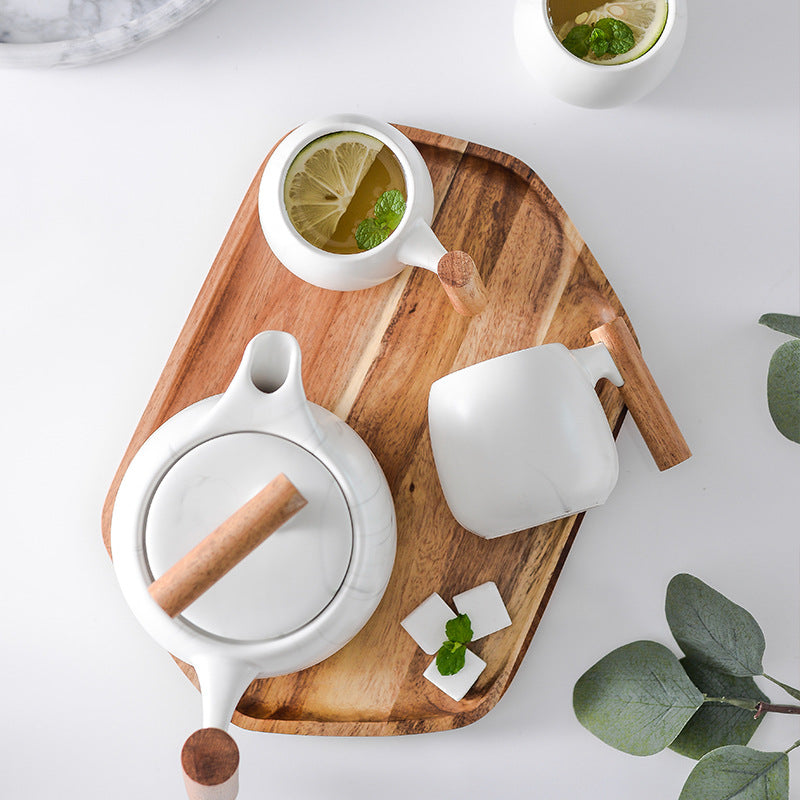 White ceramic tea set with light wood handles and a lid, arranged on a wooden tray with a cup of mint tea, tea cubes, and green leaves on a white background.