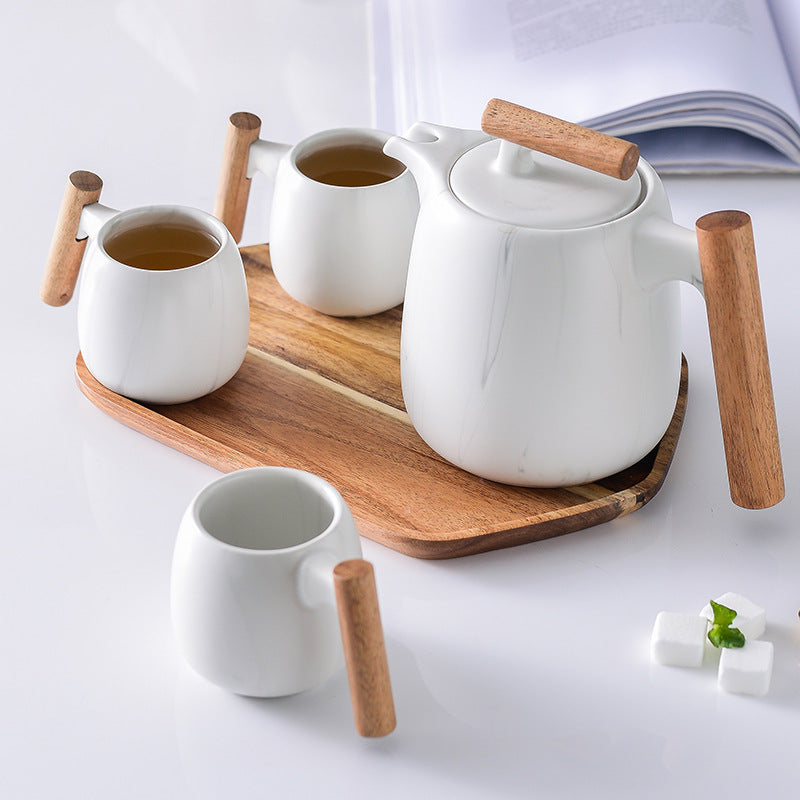 White ceramic tea set with wooden handles and a matching wooden tray, including a teapot, teacups, and a small cup, arranged on a white surface with sugar cubes and an open book in the background.