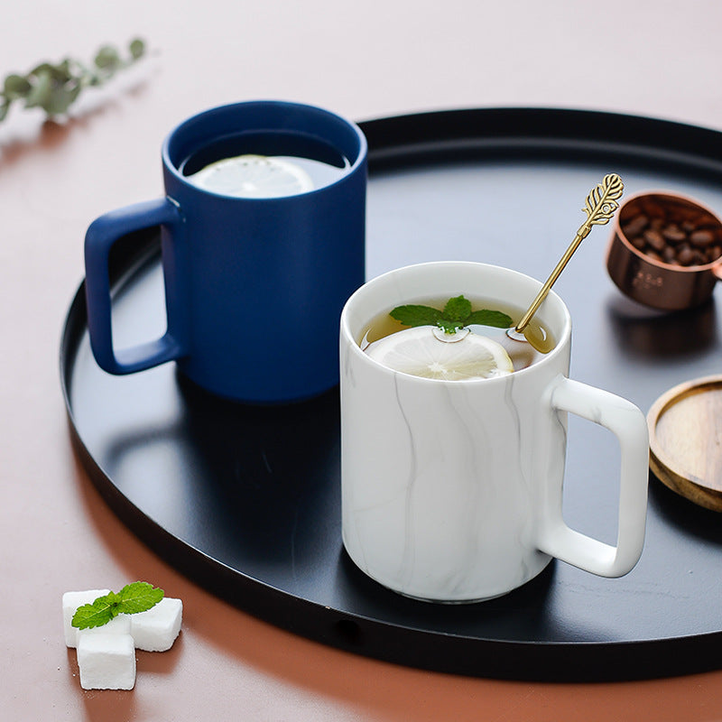 Two ceramic mugs on a black tray: one blue mug filled with milk and one white marble mug filled with tea and mint, styled on a pink surface with small props.
