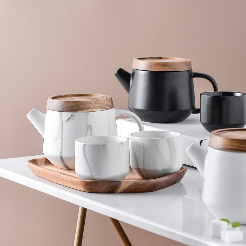 A styled tea set arrangement on a white table against a warm beige background, featuring a white marble-patterned teapot and cups with wooden lids, alongside a matte black teapot and cup on a white tray.