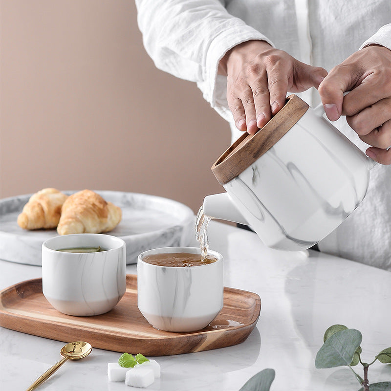 A person in a white shirt pours hot water from a white marble teapot with a wooden lid into two matching white marble cups on a wooden tray, with croissants and a spoon in the background.
