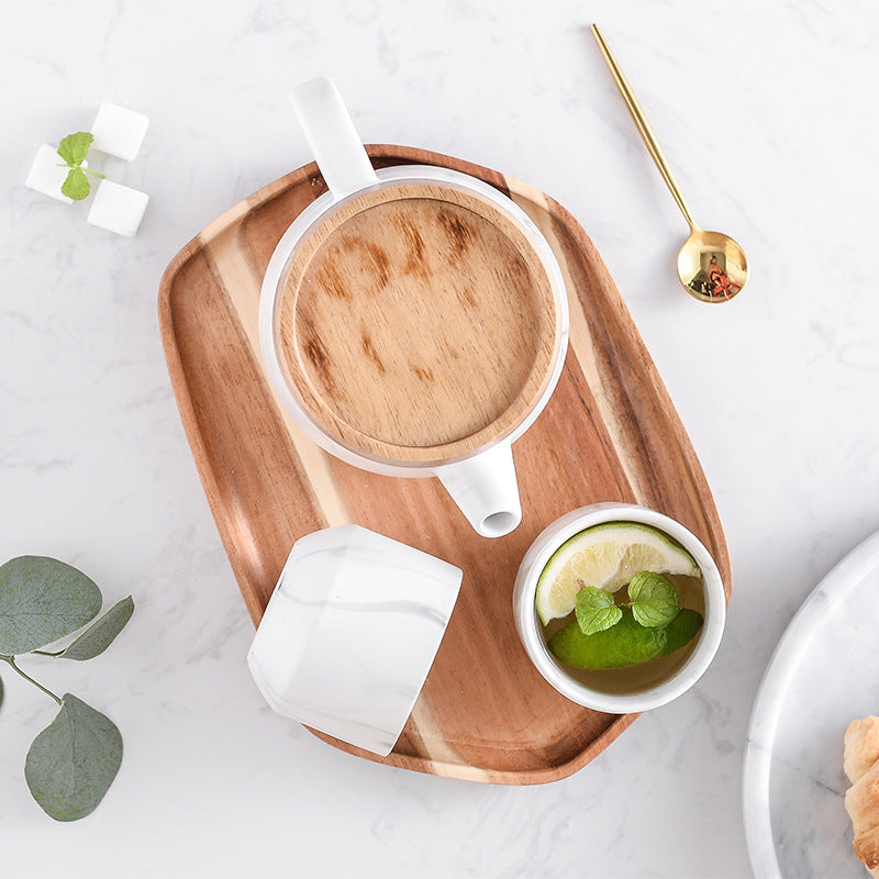 A styled overhead flat-lay of a white ceramic teapot with a light wood lid, a matching white cup, and a small white cup holding lime slices and mint, arranged on a wooden tray on a white marble surface with a gold spoon nearby.