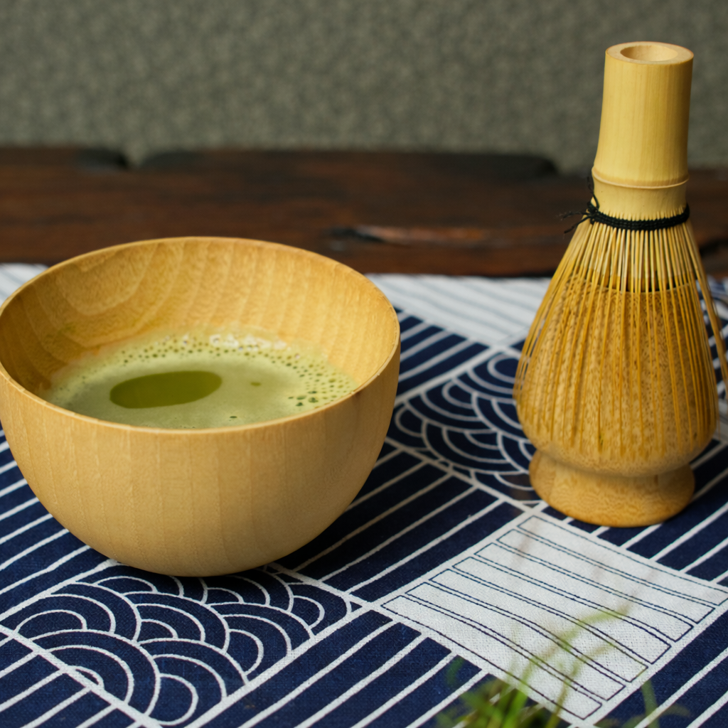 Wooden bowl with green tea and bamboo whisk on a patterned tablecloth