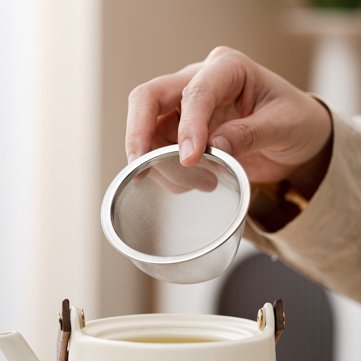 A close-up of a hand holding a small stainless steel mesh tea infuser above a white teapot, with a softly blurred indoor background.
