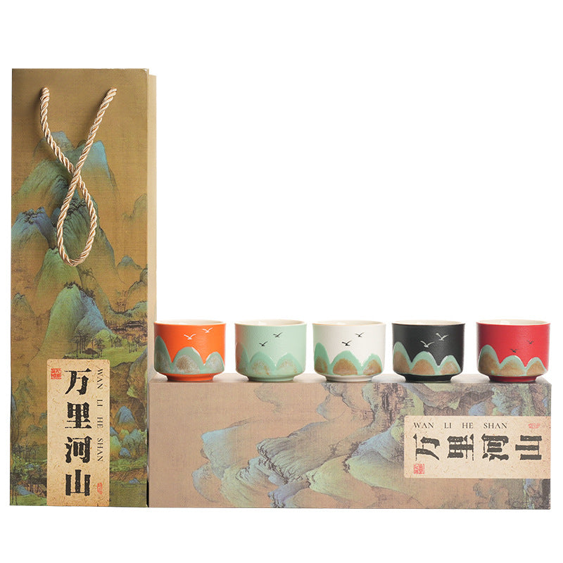 A tea gift set with five small ceramic cups in orange, mint green, white, black, and red, displayed on top of a decorative box next to a matching paper gift bag with a rope handle. The packaging features traditional Chinese landscape artwork and the text 'WAN LI HE SHAN'.