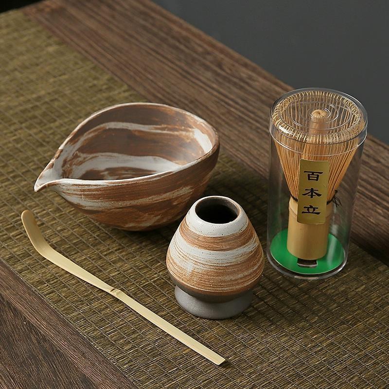 Tea-making set with ceramic bowl, whisk, scoop, and container on a wooden surface.