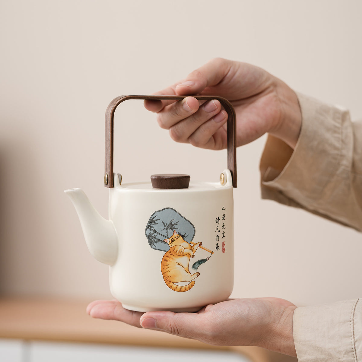 A hand-painted ceramic teapot with a white body, brown handle, and an illustration of an orange tabby cat on a gray rock, held by two hands against a soft beige background.