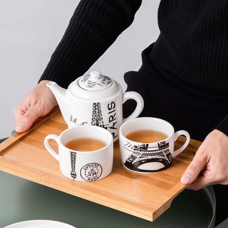 A person in a black sweater holds a bamboo tray with a white ceramic teapot and two matching cups decorated with black Eiffel Tower and Paris text designs, set against a neutral gray background.