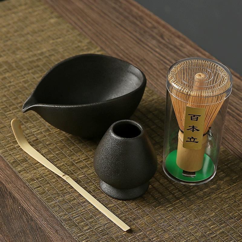 Tea-making set with black ceramic pitcher, small pot, gold whisk, and transparent container on a wooden surface.
