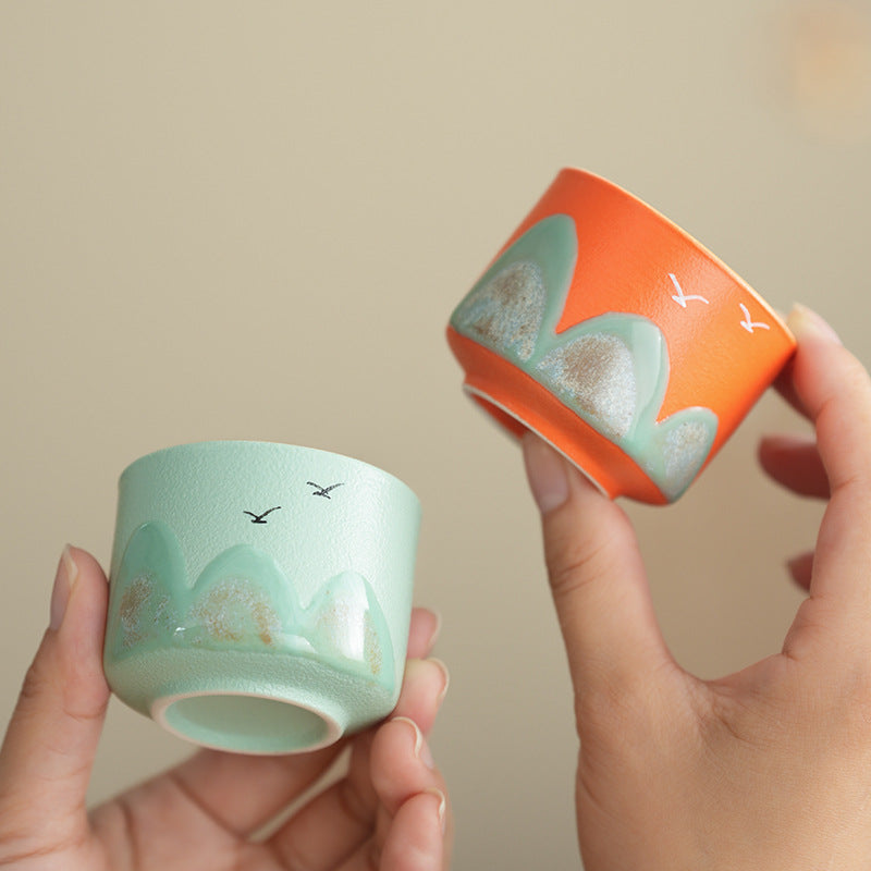 Two small ceramic cups with hand-painted mountain and bird designs, one in mint green and the other in orange, held in two hands against a soft beige background.