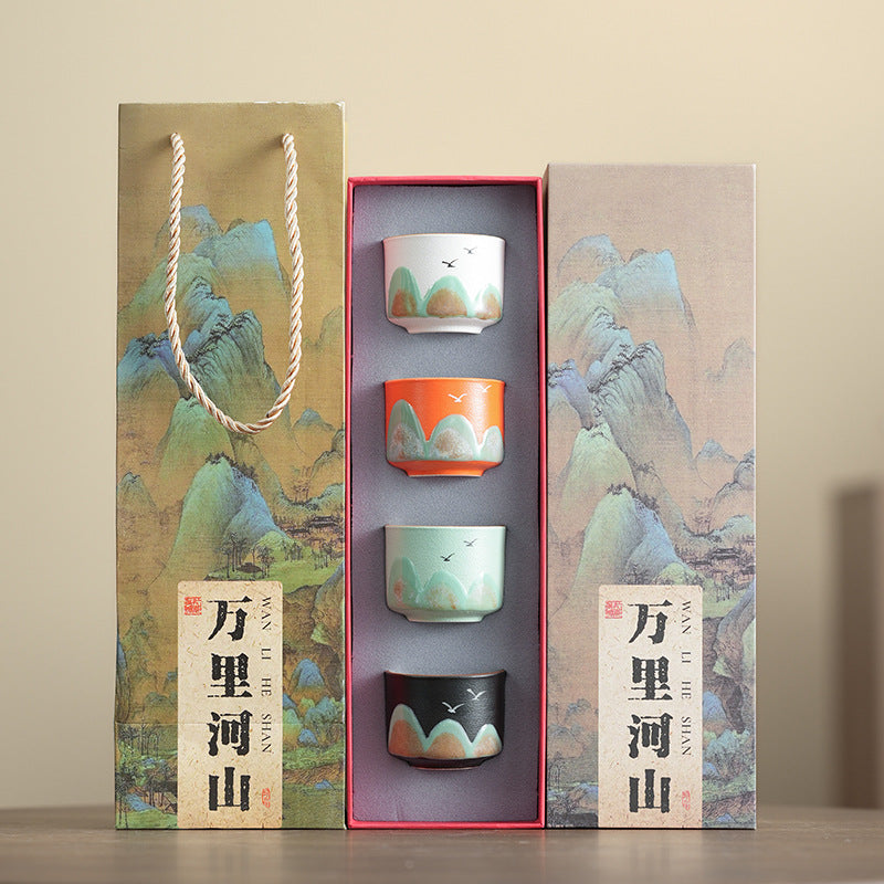 A ceramic tea set gift set with four small cups in white, orange, mint green, and black, decorated with traditional Chinese mountain landscape paintings, shown with its gift packaging.