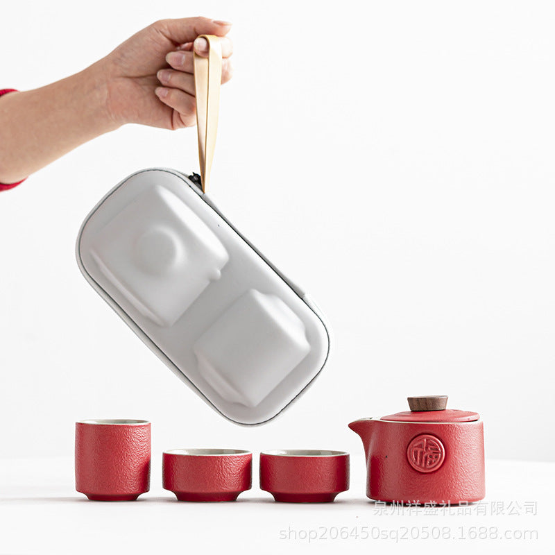 A hand holds a silver metal tea bag holder above a red ceramic teapot and four matching red cups on a white background.