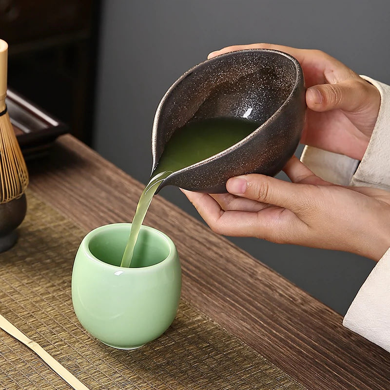 Person pouring green liquid from a ceramic pitcher into a green cup on a wooden table.