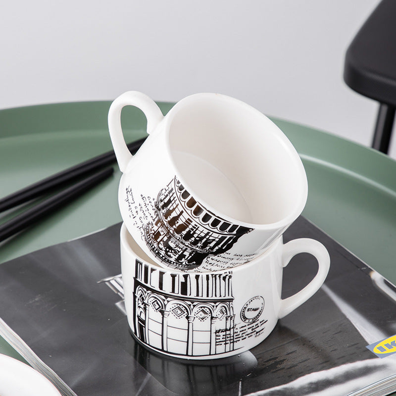 Two stacked white ceramic coffee mugs with black architectural illustrations, displayed on a glossy magazine over a green tray.