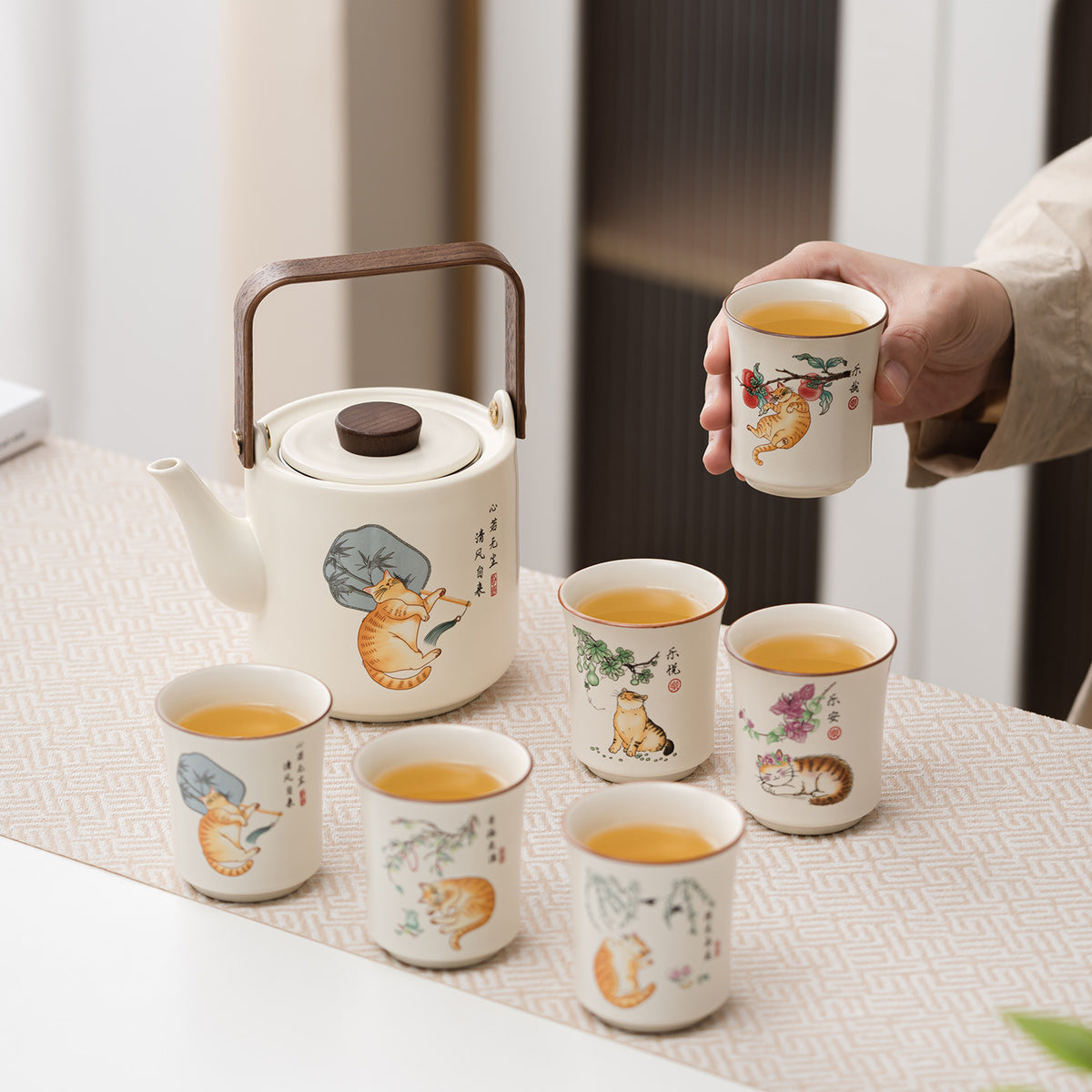 A ceramic tea set with a white teapot and five matching cups decorated with colorful cat illustrations, arranged on a table runner. A hand is holding one of the cups filled with tea, and the background shows a softly blurred indoor setting.