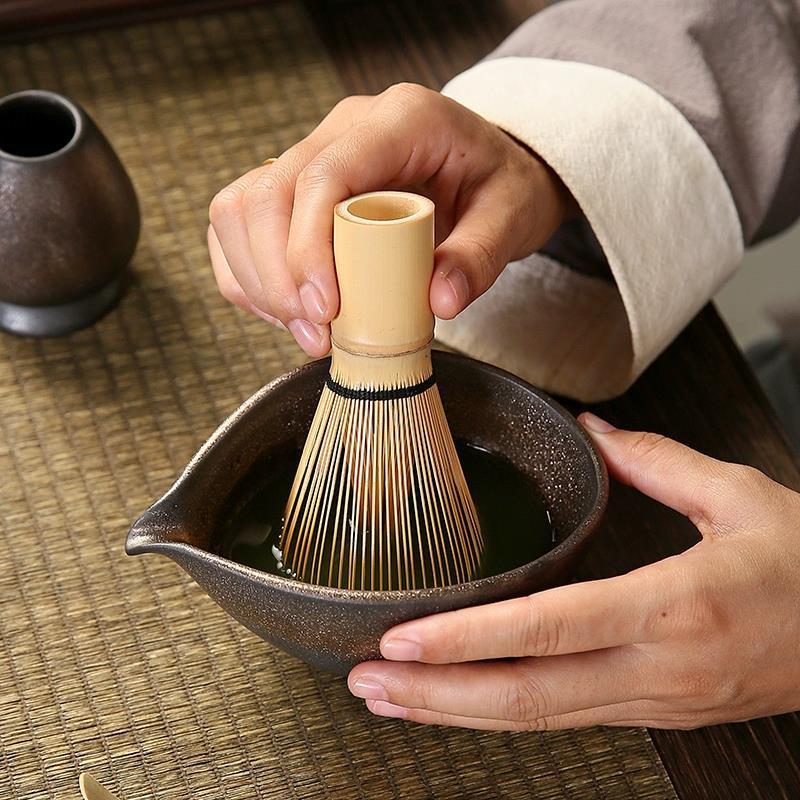 Person using a bamboo whisk in a dark ceramic bowl on a wooden surface
