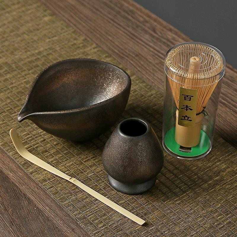 Tea-making set with black ceramic pitcher, small pot, gold spoon, and bamboo whisk on a wooden surface.