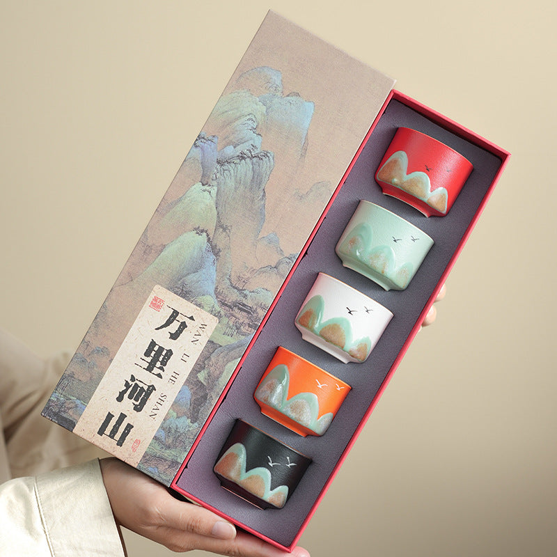 A hand holds an open presentation box containing four small ceramic cups with matching landscape-style prints. The box has a beige illustrated cover with Chinese text and a red inner tray holding the cups.