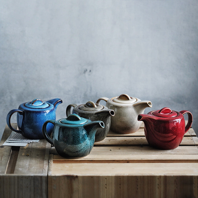 Five ceramic teapots in assorted colors including blue, teal, gray, beige, and red, arranged on a rustic wooden table against a textured gray wall.