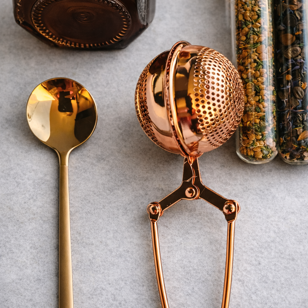 Rose gold tea infuser and spoon on a light gray surface with tea leaves in the background.